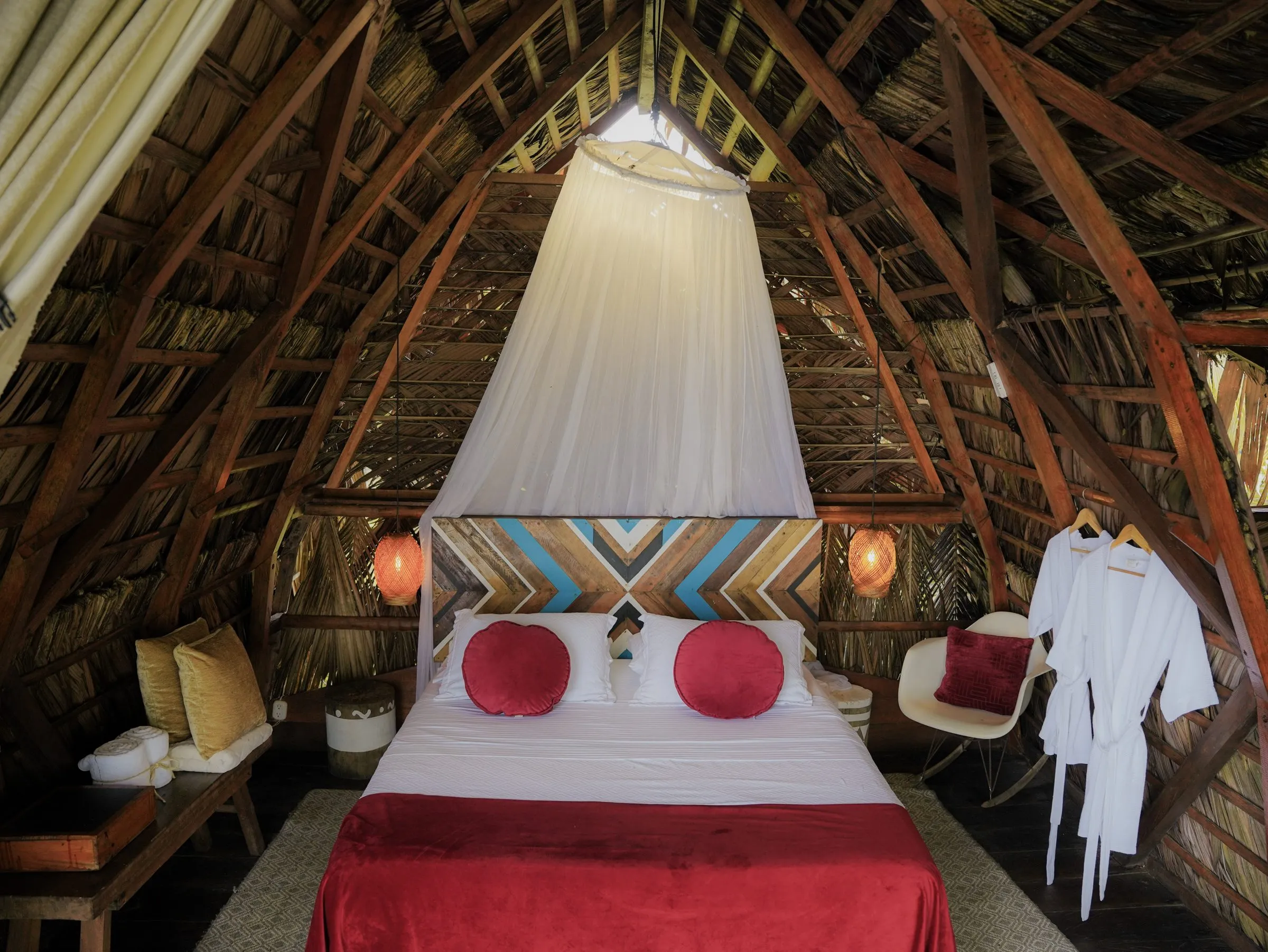 Cama del Bungalow Maxi 1 con mosquitero, cojines rojos y batas colgadas bajo el techo de palma